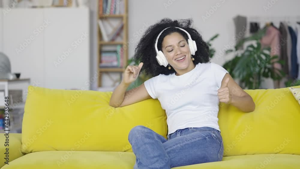 Portrait of a young African American woman enjoys music with big white headphones. Brunette with curly hair sitting on yellow sofa in a bright home room. Slow motion ready 59.97fps. Portrait of a young African American woman enjoys music with big white headphones. Brunette with curly hair sitting on yellow sofa in a bright home room. Slow motion ready 59.97fps.