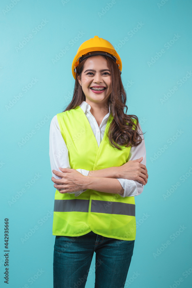 Confident beautiful young engineer woman smiling in yellow hard hat with arms crossed isolated on mint blue background. Confident beautiful young engineer woman smiling in yellow hard hat with arms crossed isolated on mint blue background.