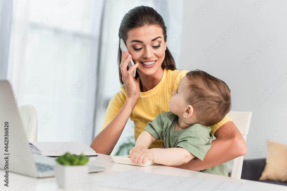 Smiling mother talking on smartphone near toddler son with calculator Smiling mother talking on smartphone near toddler son with calculator