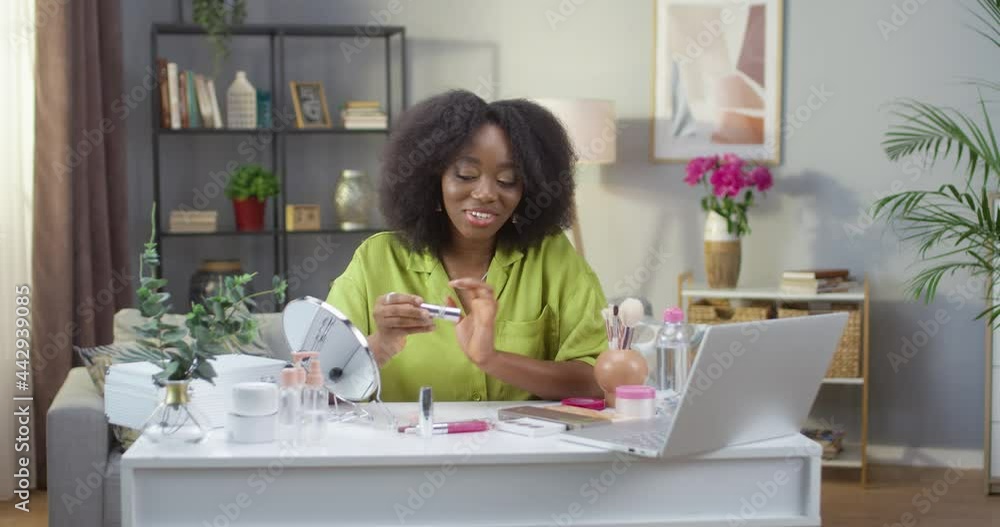 Portrait of happy pretty African American young woman stylist sitting at table in modern room speaking on video call on laptop showing lipstick. Beauty blogger making review on cosmetics product Portrait of happy pretty African American young woman stylist sitting at table in modern room speaking on video call on laptop showing lipstick. Beauty blogger making review on cosmetics product