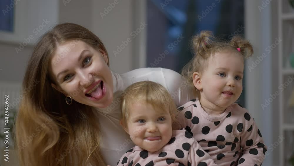 Charming baby twins and mother having fun showing tongues posing indoors. Portrait of relaxed Caucasian woman and girls in living room at home. Family concept Charming baby twins and mother having fun showing tongues posing indoors. Portrait of relaxed Caucasian woman and girls in living room at home. Family concept