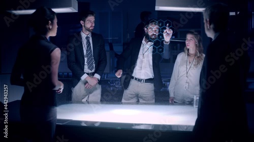 Businessman in suit talking to business people colleagues or partners standing around futuristic table with light showing an architecture project powered by a holographic modern projection technology.