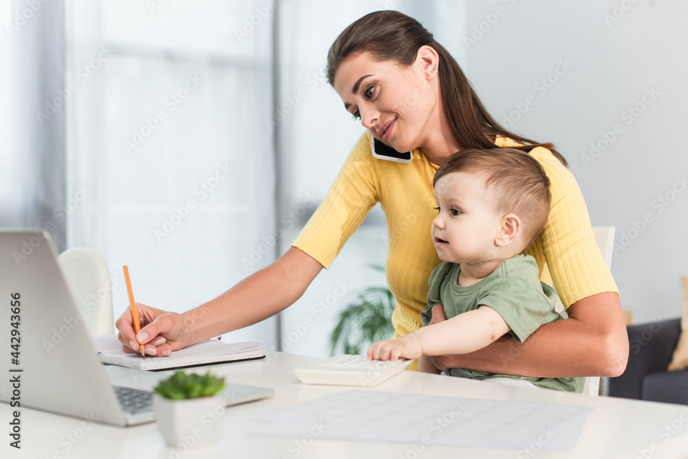 Mother talking on mobile phone while working near toddler son at home Mother talking on mobile phone while working near toddler son at home