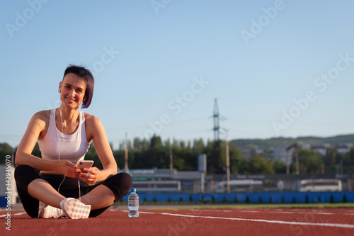 A cheerful young woman in sportswear relaxing after a workout or run, sitting on a stadium track, taking selfies, listening to music on headphones on her smartphone, taking a break during a workout