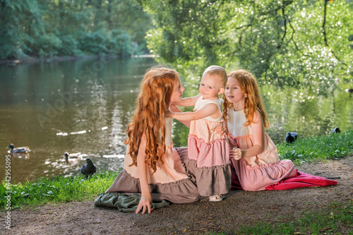 Three red-haired sisters in long linen dresses blow bubbles in the park on sunny summer day. © Maria Moroz