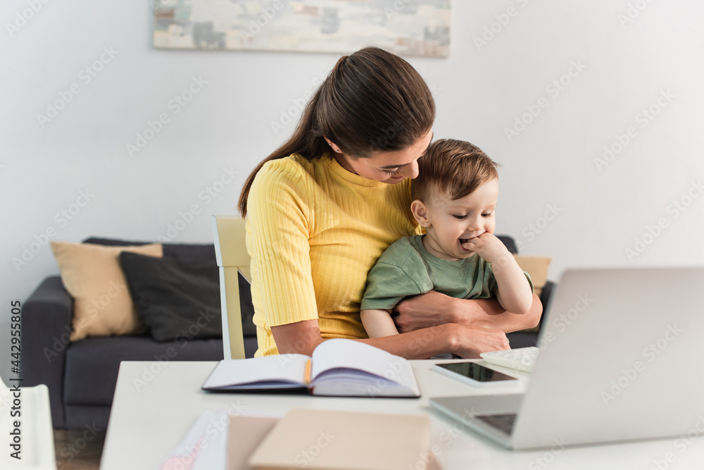 Young mother embracing toddler kid near gadgets and notebooks on blurred foreground Young mother embracing toddler kid near gadgets and notebooks on blurred foreground