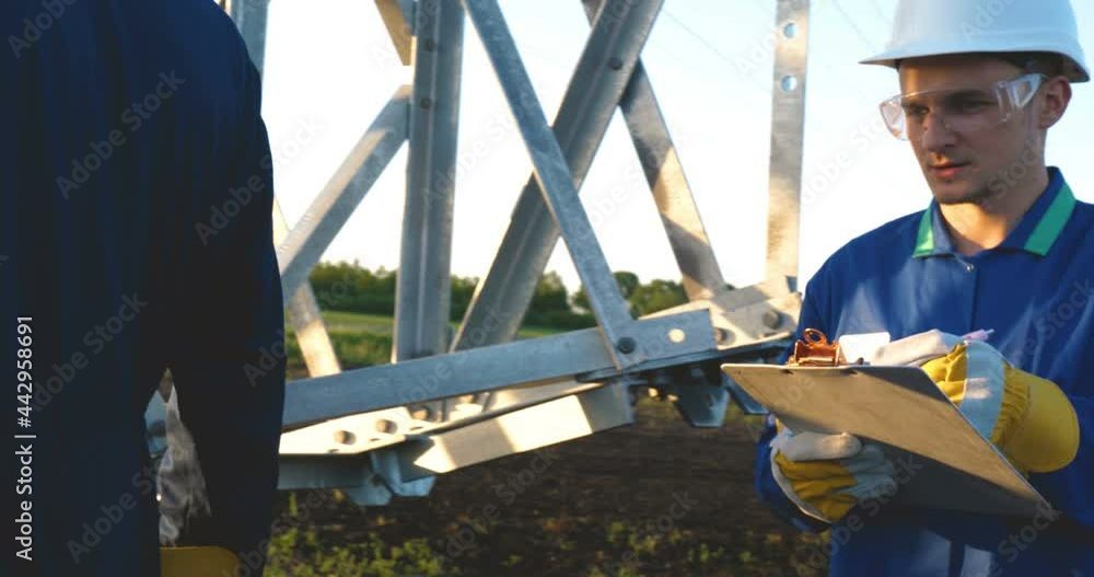 An electrician or builder and engineer, in a blue robe, in a yellow and white helmet, check the technical overview for strength, prepare electric poles, an electric tower, energy, and workers. An electrician or builder and engineer, in a blue robe, in a yellow and white helmet, check the technical overview for strength, prepare electric poles, an electric tower, energy, and workers.
