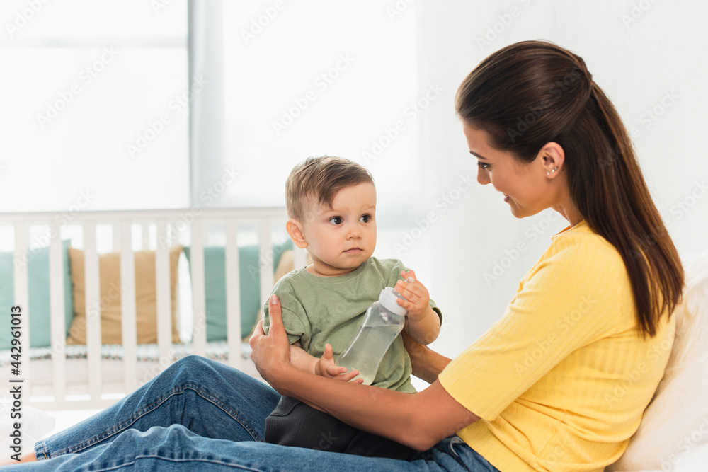 Smiling mother hugging son with baby bottle on bed Smiling mother hugging son with baby bottle on bed