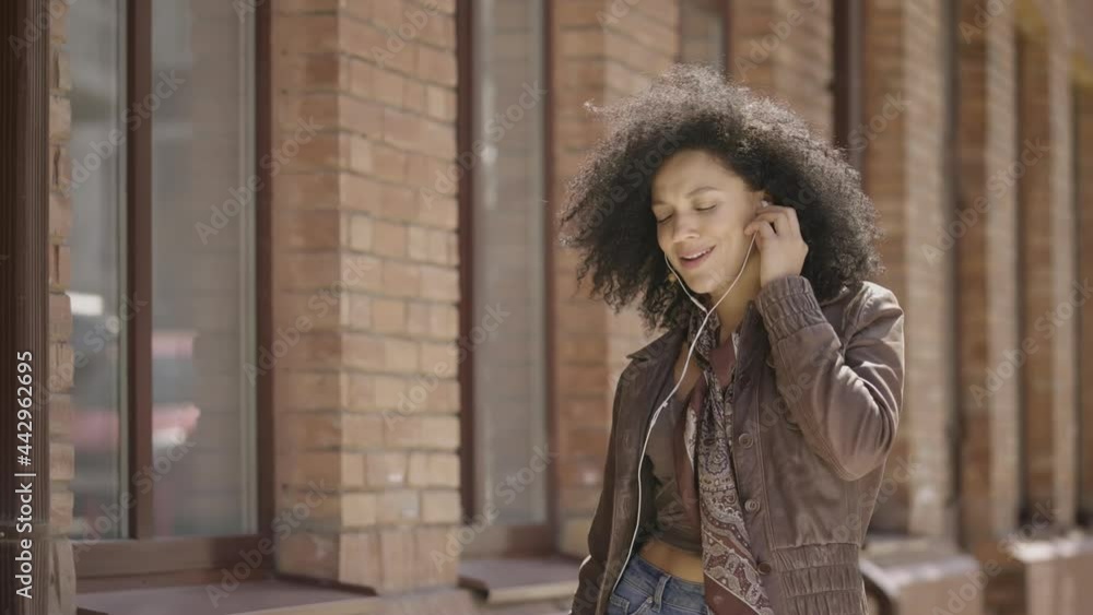 Portrait of young African American woman dancing and enjoying music with headphones using phone. Brunette in leather jacket walks street against blurred building. Close up. Slow motion ready 59.97fps. Portrait of young African American woman dancing and enjoying music with headphones using phone. Brunette in leather jacket walks street against blurred building. Close up. Slow motion ready 59.97fps.