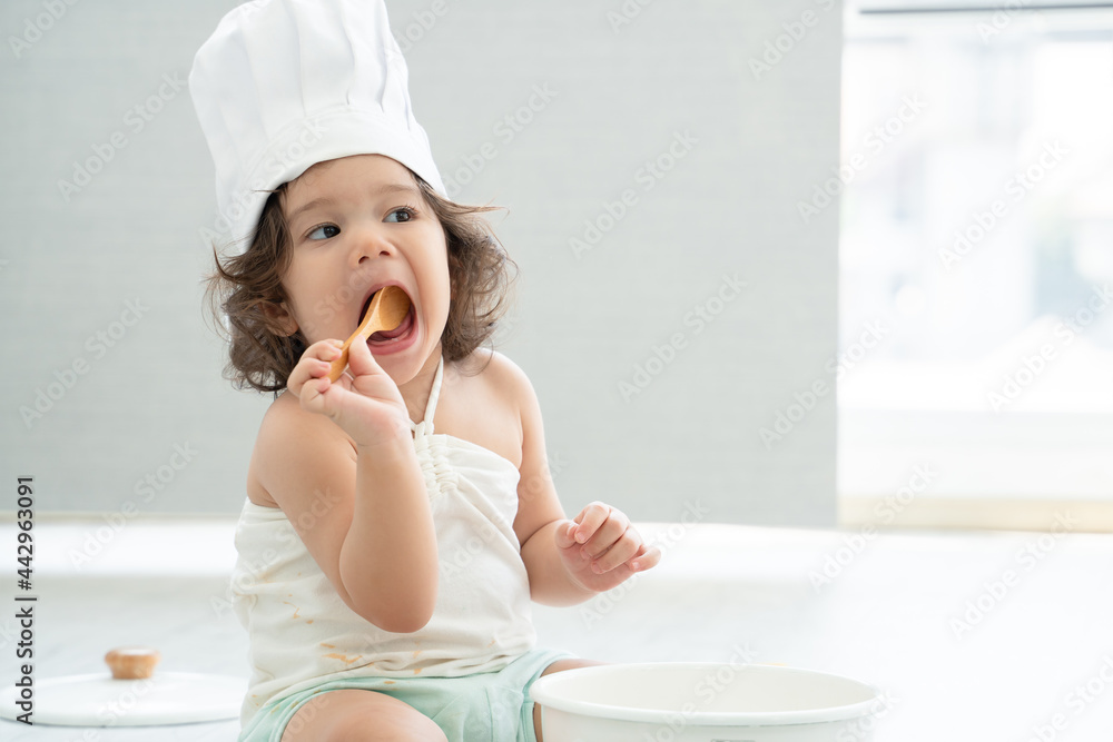 Portrait of little Caucasian adorable kid girl wear chef hat holding wooden spoon and tasting food from pot with white t-shirt mess up with ketchup. Cute child sitting on floor at kitchen at home. Portrait of little Caucasian adorable kid girl wear chef hat holding wooden spoon and tasting food from pot with white t-shirt mess up with ketchup. Cute child sitting on floor at kitchen at home.