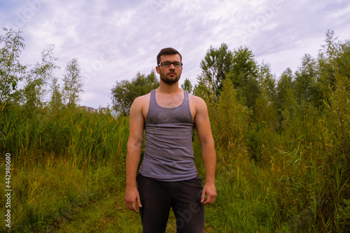 A young man in gray clothes and glasses is standing in nature near green bushes. Sports tourism in nature