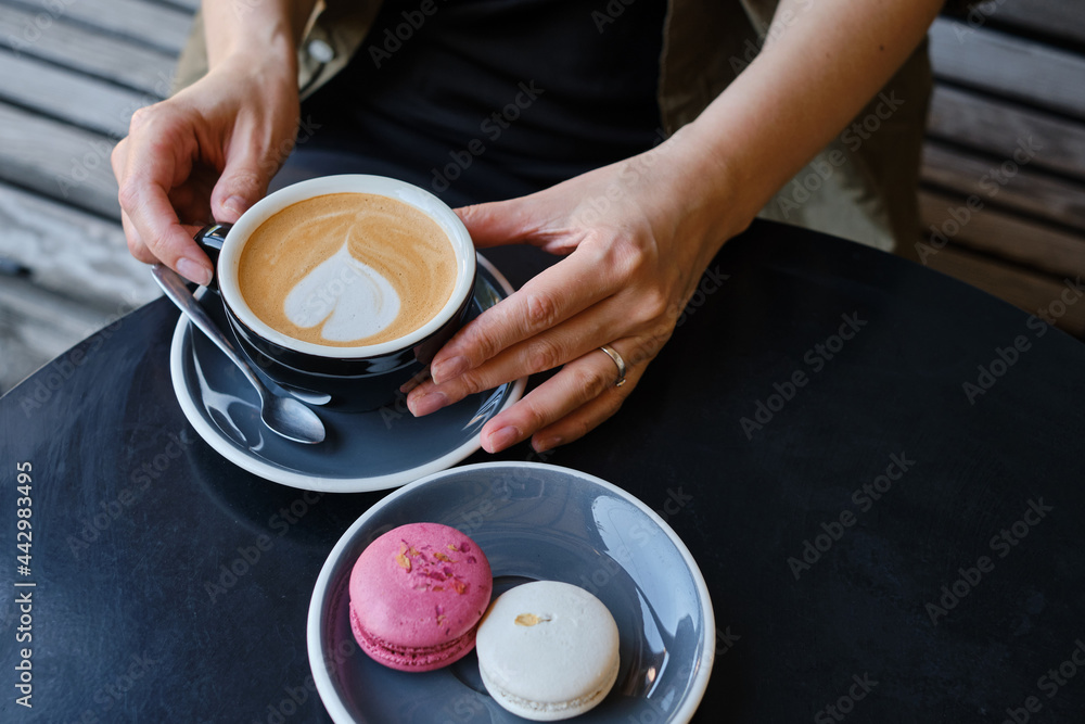 Woman with a cup of aromatic coffee in her hands sits on the summer terrace of the restaurant. The girl drinks hot latte after breakfast with sweet macaroon dessert. Delicious French dessert macaroon