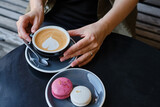 Woman with a cup of aromatic coffee in her hands sits on the summer terrace of the restaurant. The girl drinks hot latte after breakfast with sweet macaroon dessert. Delicious French dessert macaroon