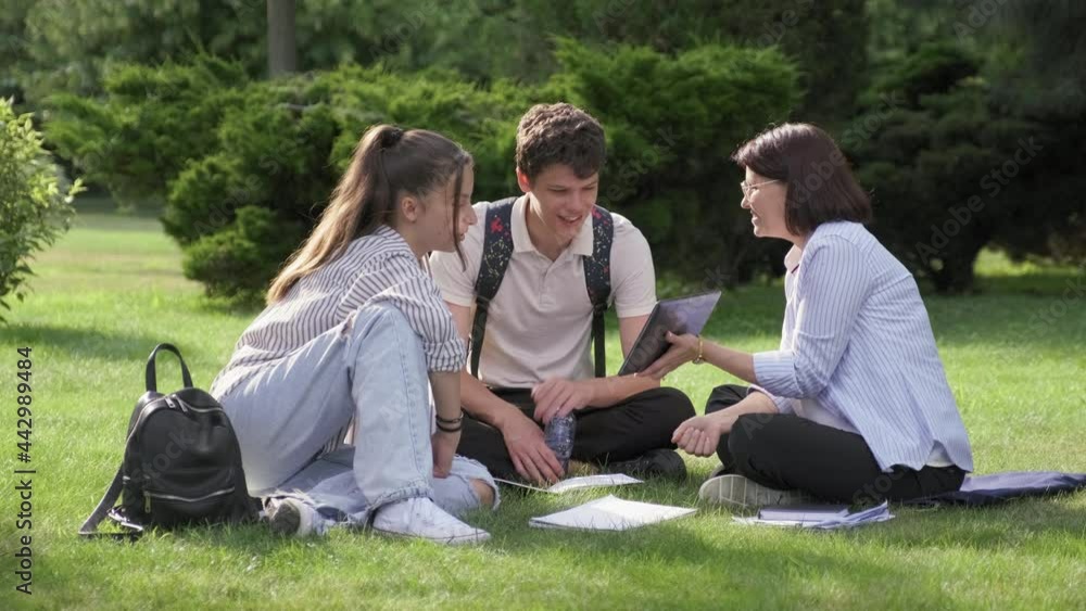 School teacher, psychologist, social worker talking to teenagers, sitting on grass School teacher, psychologist, social worker talking to teenagers, sitting on grass