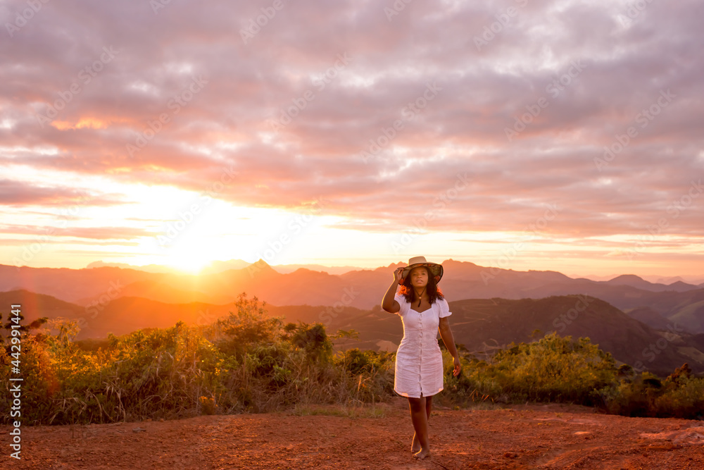 Young woman barefoot with arms raised and wearing a hat, enjoying nature, with the sunset in the background.
