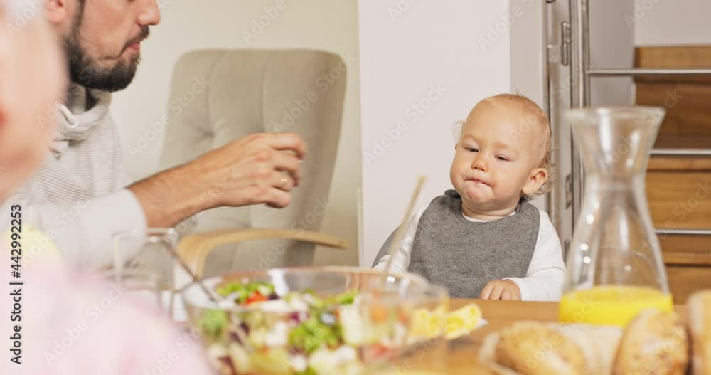 Young dad, smiling men, sits next to child at feeding chair, feeds son with roll, bread, boy bites food with appetite, satisfied, in front of them richly set table, juice, salad, guy talks to toddler Young dad, smiling men, sits next to child at feeding chair, feeds son with roll, bread, boy bites food with appetite, satisfied, in front of them richly set table, juice, salad, guy talks to toddler