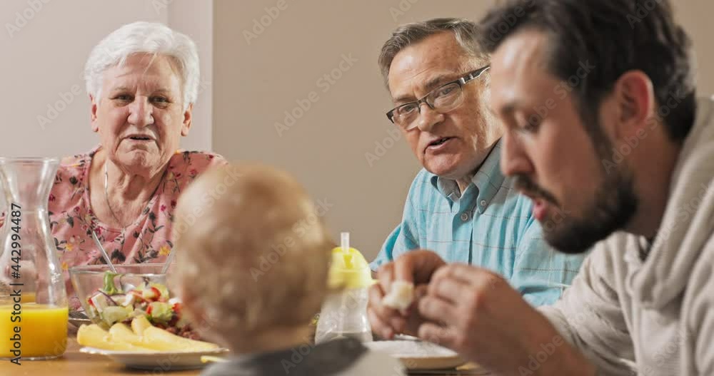 A brunette men sits at the table with his parents and feeds his little, cute, sweet son with blond hair, grandparents admire their grandson's appetite, they eat breakfast, family atmosphere A brunette men sits at the table with his parents and feeds his little, cute, sweet son with blond hair, grandparents admire their grandson's appetite, they eat breakfast, family atmosphere
