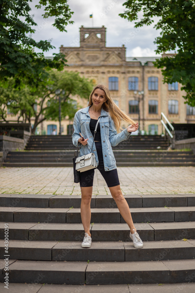 Young beautiful blonde woman in a denim jacket and tight sports shorts stands on the stairs