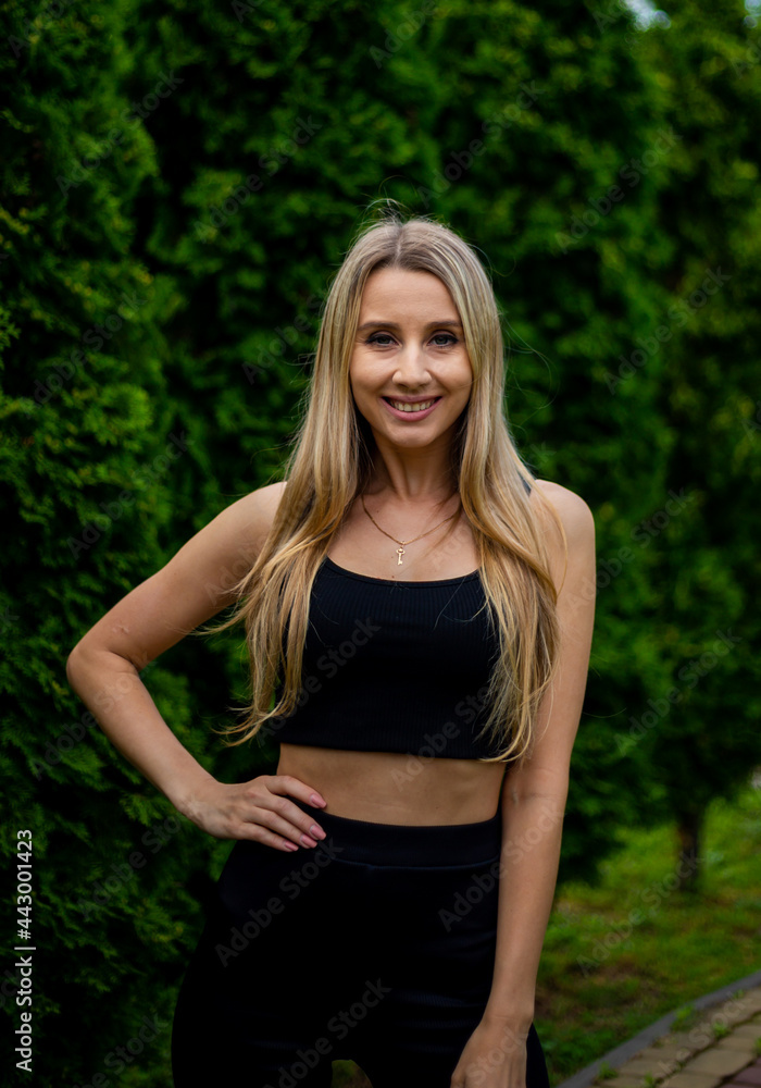 Young beautiful sporty blonde woman in a black T-shirt and in black tight sports shorts in good shape posing in front of green trees Young beautiful sporty blonde woman in a black T-shirt and in black tight sports shorts in good shape posing in front of green trees