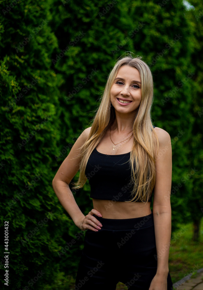 Young beautiful sporty blonde woman in a black T-shirt and in black tight sports shorts in good shape posing in front of green trees Young beautiful sporty blonde woman in a black T-shirt and in black tight sports shorts in good shape posing in front of green trees