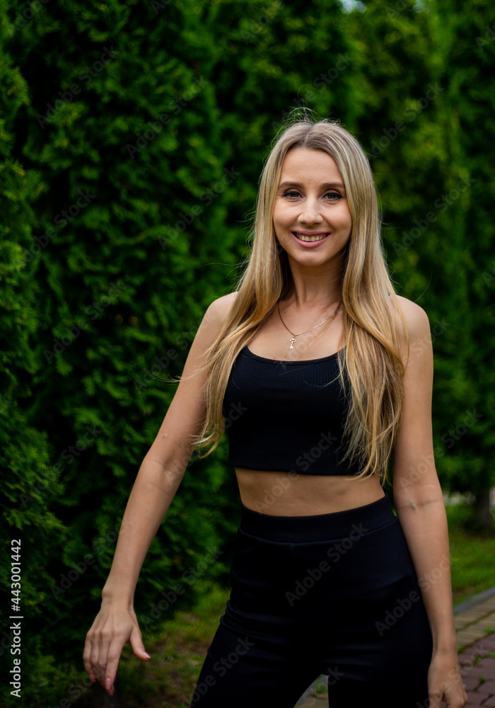 Young beautiful sporty blonde woman in a black T-shirt and in black tight sports shorts in good shape posing in front of green trees Young beautiful sporty blonde woman in a black T-shirt and in black tight sports shorts in good shape posing in front of green trees