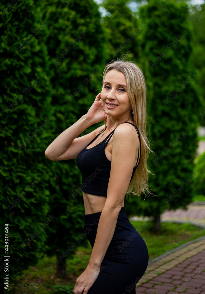 Young beautiful sporty blonde woman in a black T-shirt and in black tight sports shorts in good shape posing in front of green trees