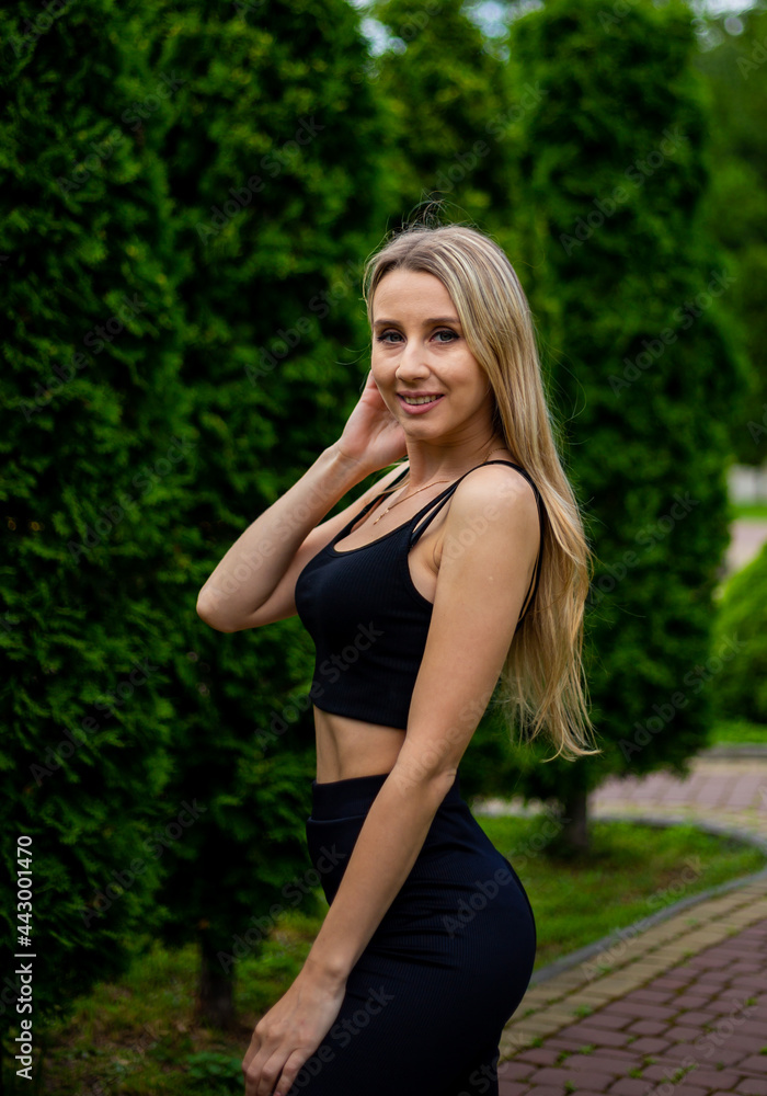Young beautiful sporty blonde woman in a black T-shirt and in black tight sports shorts in good shape posing in front of green trees Young beautiful sporty blonde woman in a black T-shirt and in black tight sports shorts in good shape posing in front of green trees