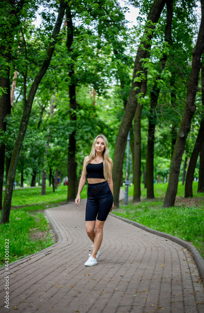 Young beautiful sporty blonde woman in a black T-shirt and in black tight sports shorts in good shape posing in front of green trees Young beautiful sporty blonde woman in a black T-shirt and in black tight sports shorts in good shape posing in front of green trees