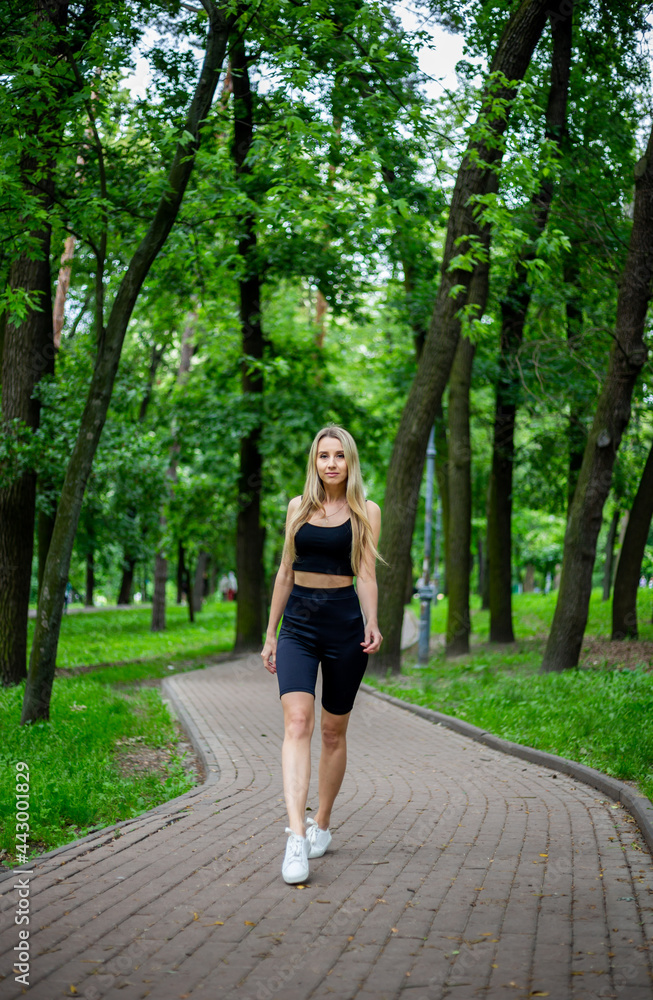 Young beautiful sporty blonde woman in a black T-shirt and in black tight sports shorts in good shape posing in front of green trees Young beautiful sporty blonde woman in a black T-shirt and in black tight sports shorts in good shape posing in front of green trees