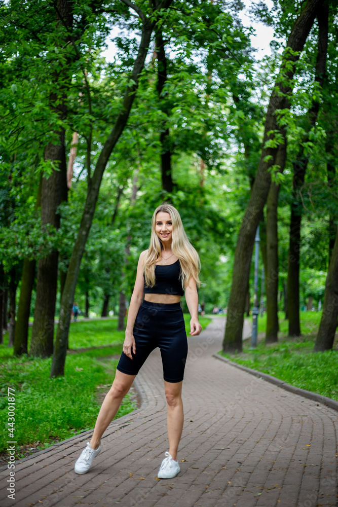 Young beautiful sporty blonde woman in a black T-shirt and in black tight sports shorts in good shape posing in front of green trees