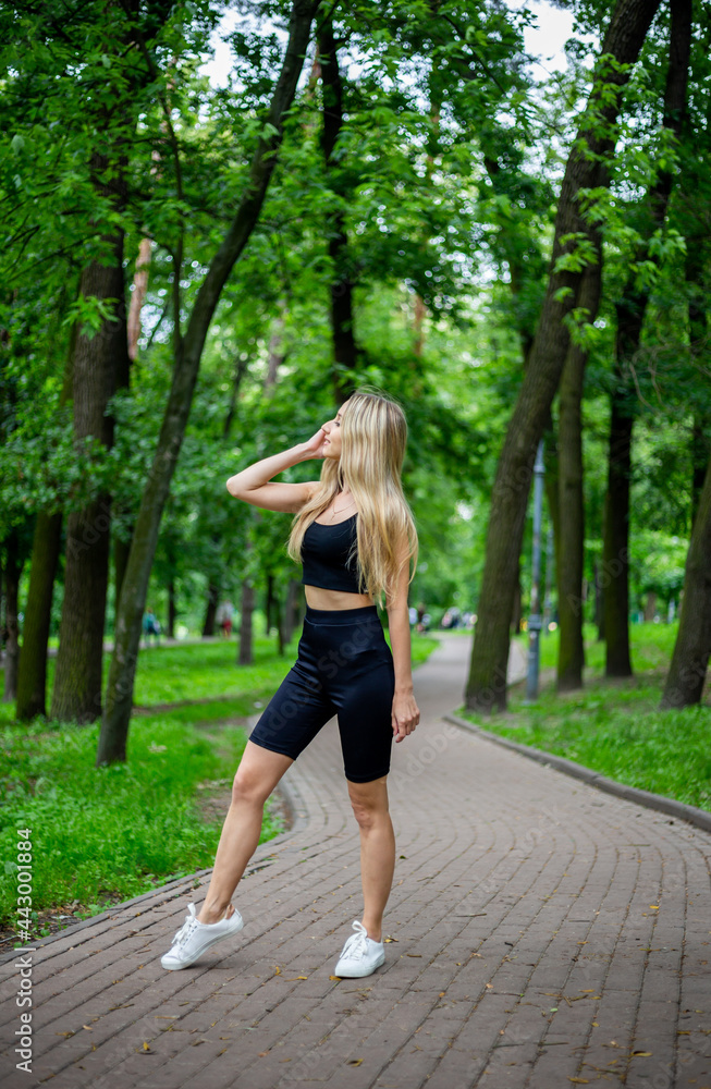 Young beautiful sporty blonde woman in a black T-shirt and in black tight sports shorts in good shape posing in front of green trees Young beautiful sporty blonde woman in a black T-shirt and in black tight sports shorts in good shape posing in front of green trees