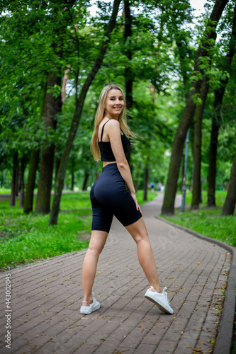 Fototapeta Naklejka Na Ścianę i Meble -  Young beautiful sporty blonde woman in a black T-shirt and in black tight sports shorts in good shape posing in front of green trees