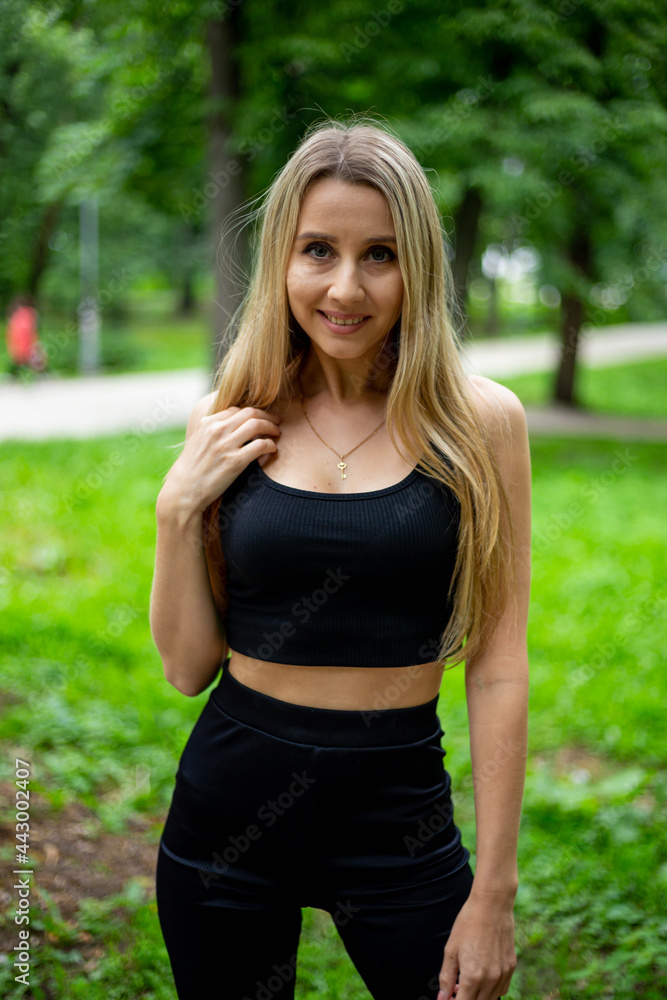 Young beautiful sporty blonde woman in a black T-shirt and in black tight sports shorts in good shape posing in front of green trees Young beautiful sporty blonde woman in a black T-shirt and in black tight sports shorts in good shape posing in front of green trees