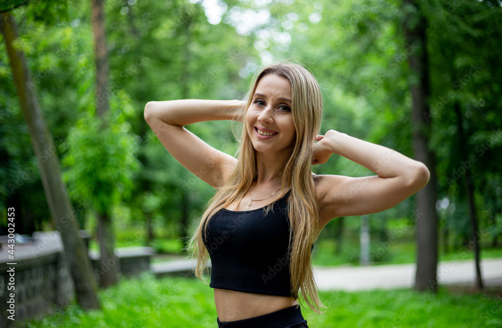 Young beautiful sporty blonde woman in a black T-shirt and in black tight sports shorts in good shape posing in front of green trees Young beautiful sporty blonde woman in a black T-shirt and in black tight sports shorts in good shape posing in front of green trees