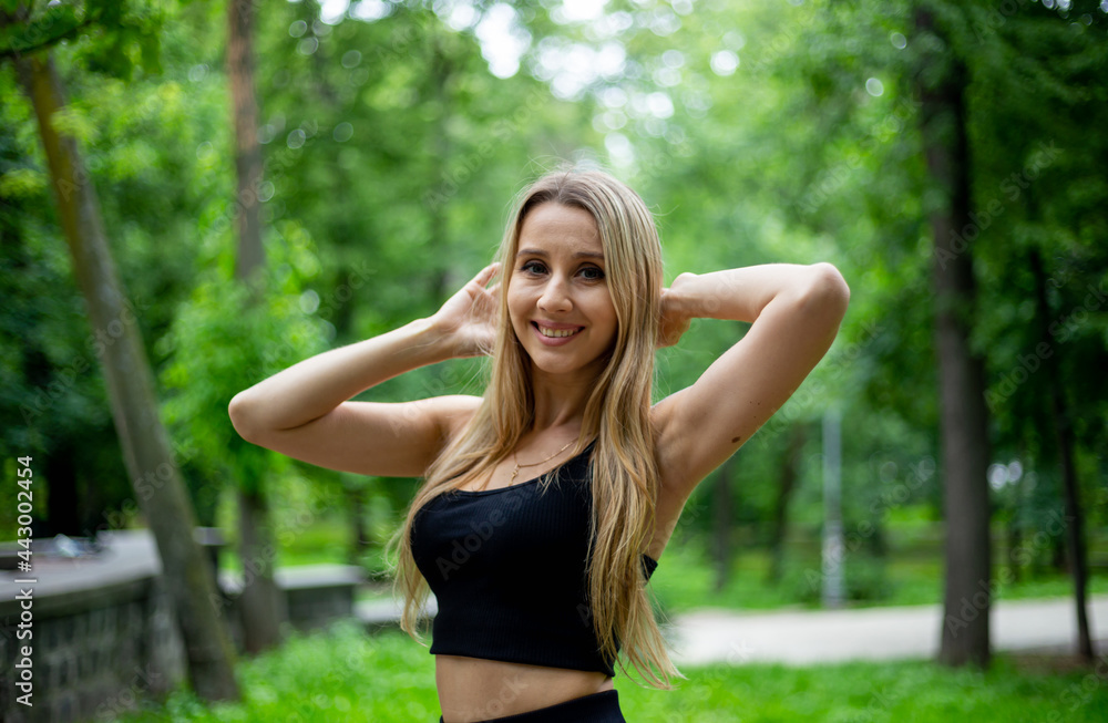 Young beautiful sporty blonde woman in a black T-shirt and in black tight sports shorts in good shape posing in front of green trees Young beautiful sporty blonde woman in a black T-shirt and in black tight sports shorts in good shape posing in front of green trees