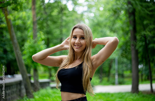 Young beautiful sporty blonde woman in a black T-shirt and in black tight sports shorts in good shape posing in front of green trees © Hennadii