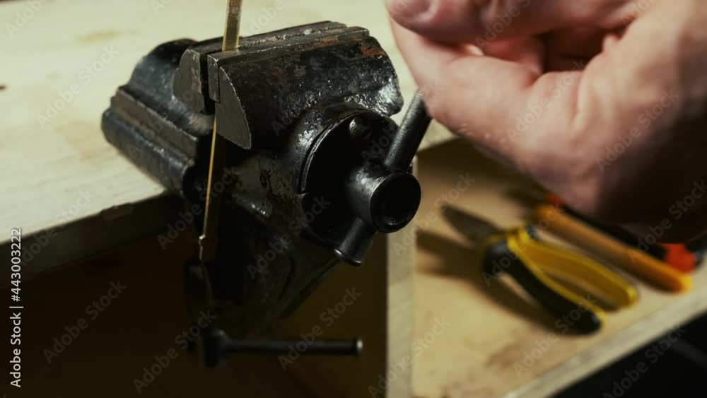 Man works with a metal iron workpiece gripped in the vise on workbench table in garage workshop. a billet is being clamped close-up to process it with a file rasp or bend it in a certain shape Man works with a metal iron workpiece gripped in the vise on workbench table in garage workshop. a billet is being clamped close-up to process it with a file rasp or bend it in a certain shape