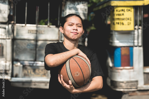 A young tomboyish woman with a smug face holds a basketball at a makeshift court where jeepneys park. photo