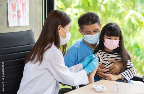 Asian  senior doctor wearing gloves and isolation mask is making a COVID-19 vaccination in the shoulder of child patient with her mother at hospital.
