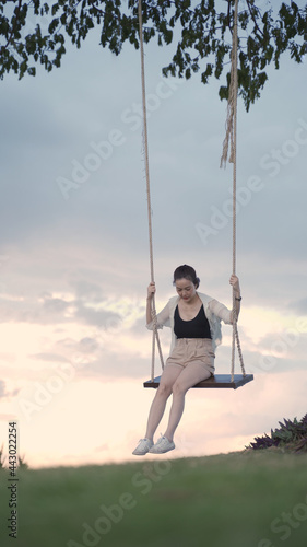 attractive asian woman tourist happy playing swing under big tree in grass field. portrait smile cheerful female swinging alone in golden time at sunset. relaxation leisure activity. copy space.