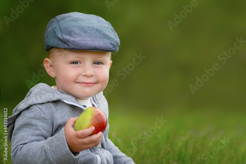 Boy in cap with apple in hand looking at camera. Blurred background. Copy space.