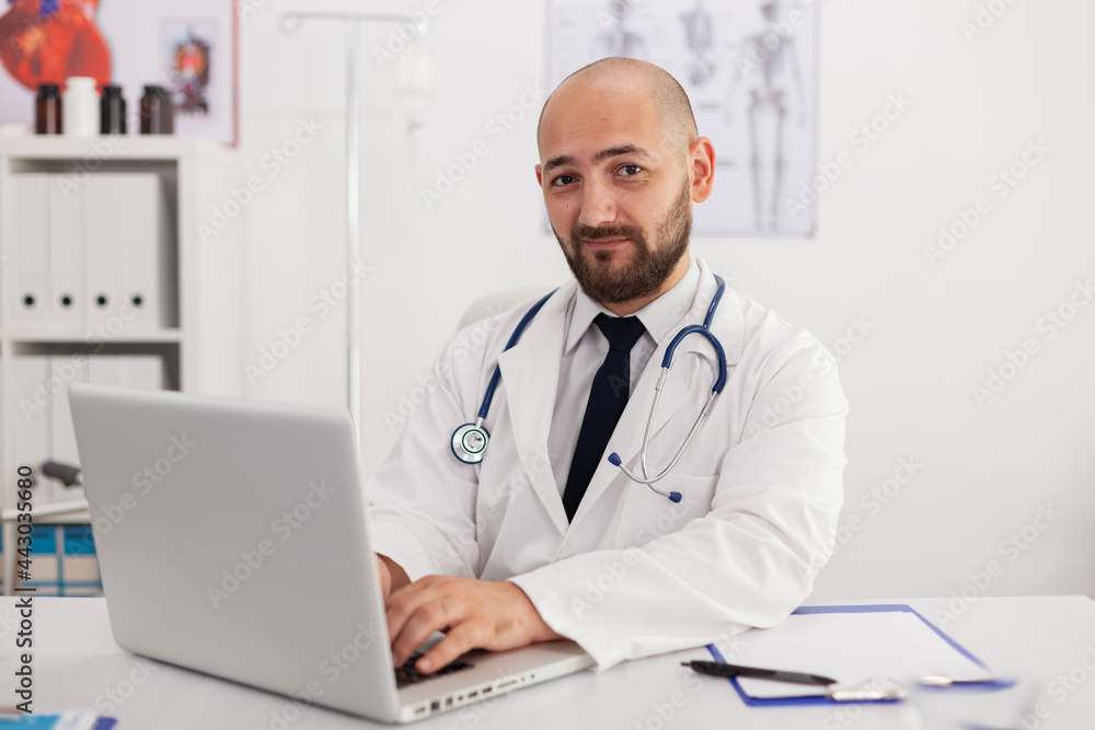 Portrait of specialist physician man looking into camera working in meeting conference room analyzing sickness expertise using laptop computer. Doctor prescribing pills medication healthcare treatment Portrait of specialist physician man looking into camera working in meeting conference room analyzing sickness expertise using laptop computer. Doctor prescribing pills medication healthcare treatment
