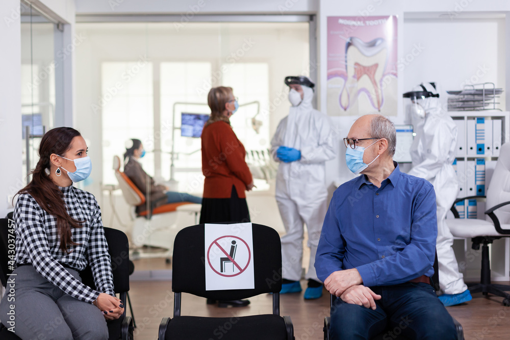 Obraz premium Senior man with face mask discussing with woman patient in stomatology clinic in waiting room, keeping social distancing during global pandemic with coronavirus. Doctor standing wearing ppe suit.