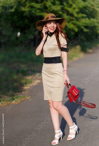 A happy woman in a hat is walking in the park in the summer with a bag and talking on the phone