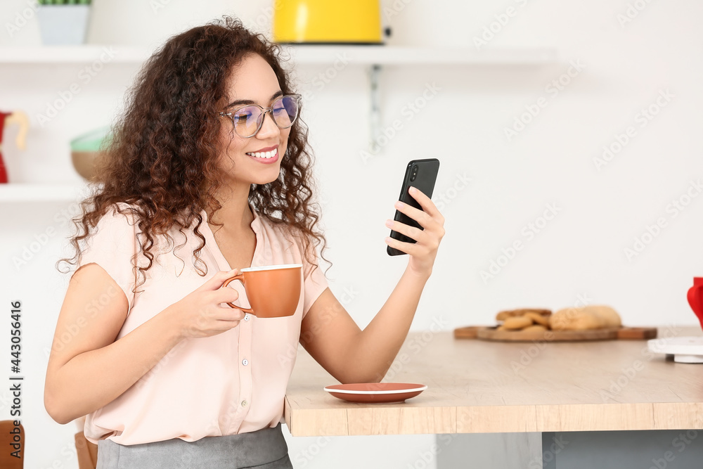 Beautiful young woman with cup of coffee using mobile phone in kitchen Beautiful young woman with cup of coffee using mobile phone in kitchen