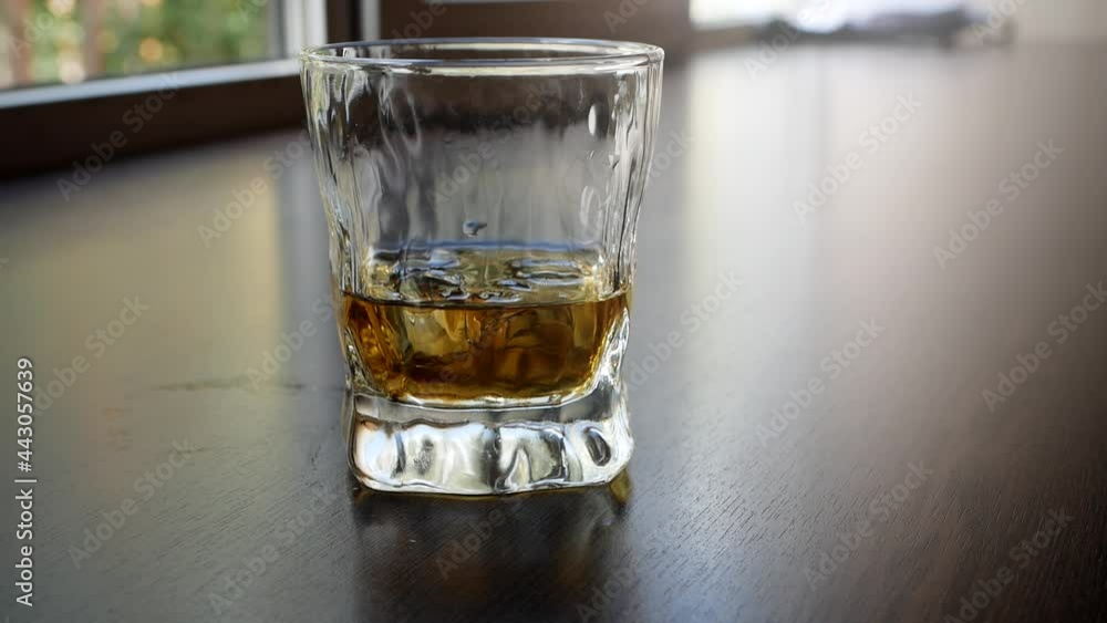 Close-up of mixing whiskey and ice cubes in a glass on a wooden table. A glass of ice and scotch whiskey. A drop of alcohol runs down the side of the glass.