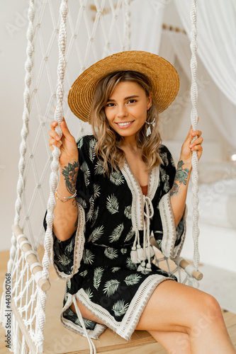 Smilling young woman in boho dress posing in stylish bedroom.  Sraw hat, boho feather drop earrings . Lady sitting on hanging swing.