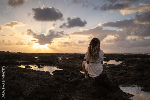 A blonde woman with curly hair and a white dress is sitting looking out to sea with a sky full of clouds and colors at sunset. Concept of peace, relaxation, tranquility, loneliness, vacation. © Alvaro