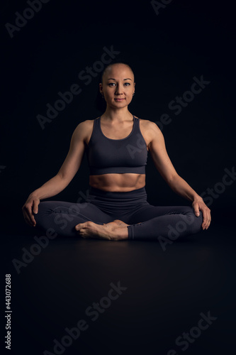 Asian woman, Fitness instructor, doing Yoga, sitting with legs crossed, in front of a black background. She looks to the camera with relaxed and confident expression.