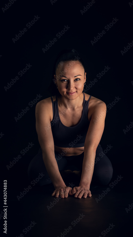 Studio photo, with black background, of an Asian woman, fitness instructor, in sports clothes, sitting on her knees, looking at the camera smiling, with a confident expression. Studio photo, with black background, of an Asian woman, fitness instructor, in sports clothes, sitting on her knees, looking at the camera smiling, with a confident expression.